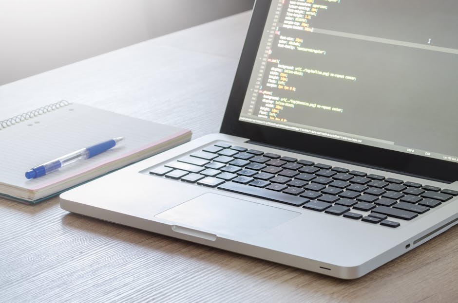 A silver laptop displaying lines of colorful code on a dark background sits open on a light wooden desk. Next to the laptop, there is an open spiral notebook with a blue and clear plastic pen resting 